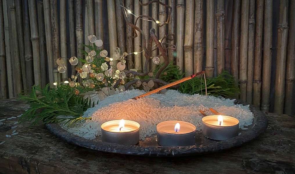 Yogic meditation Sataya -suitsuke tuoksumassa riisipedillä. Tuoksuasetelmassa on tuijan lehtiä, villiviinin oksan käppyröitä, kierrätetty lasihelminen kynttilän mansetti ja pieni tunnelmavalo tuikkukynttilöiden lisäksi.